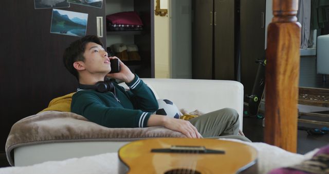 Young man sitting on couch, talking on mobile phone with relaxed expression. Headphones around neck, guitar on table nearby. Ideal for concepts of leisure, home lifestyle, technology in everyday life, modern communication, relaxation.