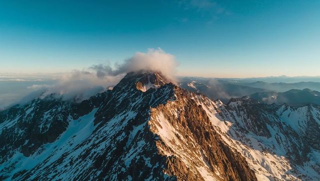Sunlit jagged mountain ridge at sunrise with cloud-capped summit and alpine snowfields panorama