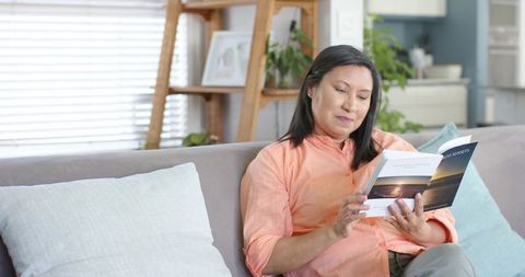 Mature woman reading paperback on cozy light gray couch in serene home interior