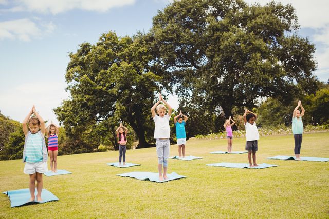 Children are practicing yoga outdoors in a park, standing on yoga mats with trees in the background. This image can be used for promoting children's fitness programs, outdoor activities, healthy lifestyle initiatives, and mindfulness practices for kids.