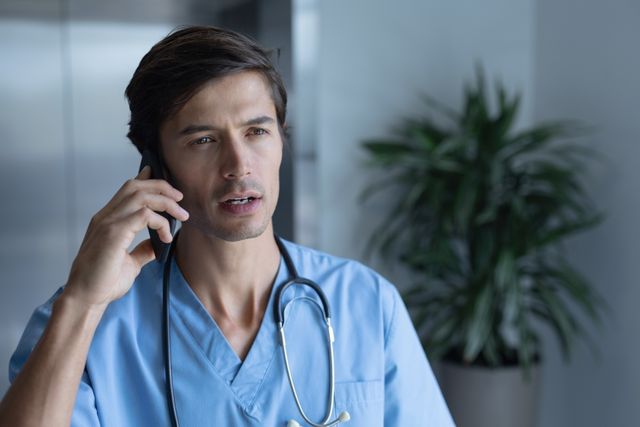 Nurse in uniform speaking on phone appears concerned, possibly discussing patient care or medical emergency. Stethoscope around neck indicates medical profession. Office background and plant create professional setting. Suitable for illustrating healthcare communication, medical services, workplace stress, telemedicine, emergencies, or hospital environment.
