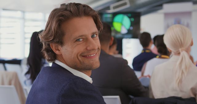 A confident businessman is smiling and looking at the camera during a presentation at a conference. In the background, other attendees are focused on a screen displaying charts. This image is ideal for showcasing business events, professional environments, leadership, and corporate teamwork.