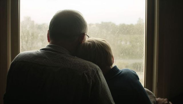 Grandfather and grandson sharing quiet moment gazing through rain-speckled window backlit