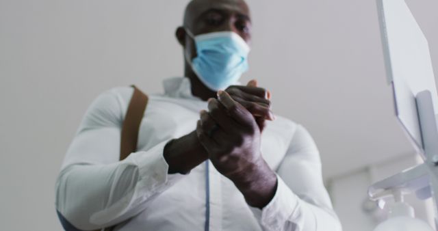 African american businessman in face mask disinfecting hands in office. business professional working in modern office during covid 19 coronavirus pandemic.