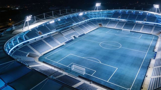 Skyview of illuminated modern soccer stadium at night with empty blue pitch and floodlights