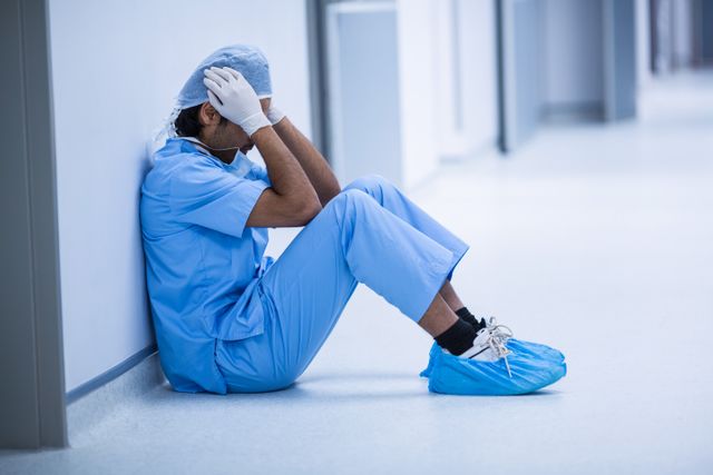 Surgeon in blue scrubs and gloves sitting on hospital corridor floor, holding head in hands, conveying stress and exhaustion. Useful for illustrating healthcare worker burnout, mental health issues in medical professionals, and the emotional toll of the medical field.