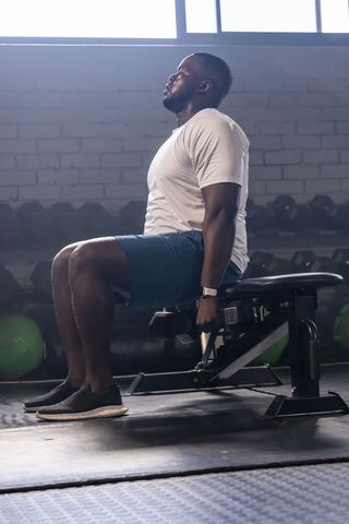 African american man holding dumbbell while seated on bench in gym showing focus