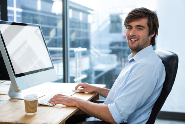 Young businessman sitting at desk in modern office, smiling at camera. Ideal for corporate websites, business presentations, and promotional materials showcasing a professional work environment.