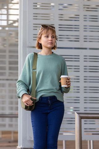 Woman commuting with coffee and crossbody bag, wearing sage sweatshirt on transit platform