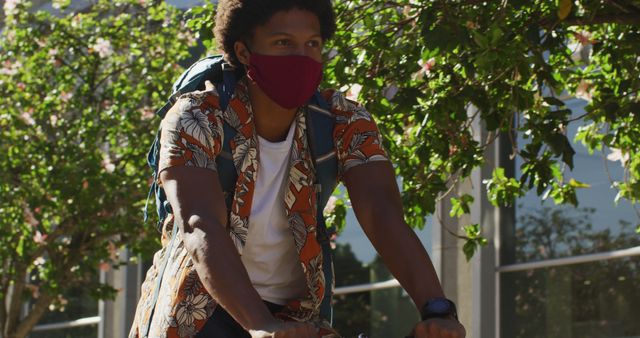 Young man riding bicycle through urban area while wearing floral shirt and face mask. Image can be used to promote safe transportation methods, healthy lifestyle, and casual fashion. Suitable for websites or publications focusing on urban living, solo activities, or outdoor exercise during global health events.
