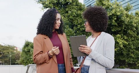 Female coworkers discussing project on rooftop terrace, holding tablet and wearing blazers