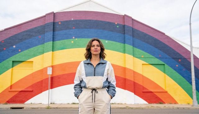 Confident woman standing in sporty windbreaker before vibrant rainbow mural on brick facade
