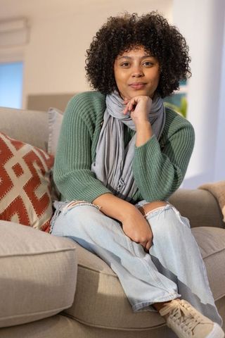 African american woman sitting on sofa wearing green sweater, relaxing in cozy living room