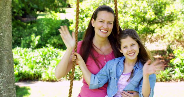 Happy mother and daughter waving while enjoying time together on a swing in a park. Ideal for use in family-related publications, parenting blogs, outdoor activity promotions, and lifestyle articles focused on relationships and nature.