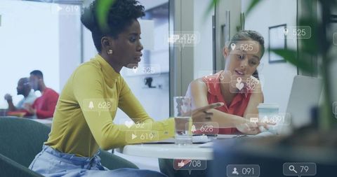 Two women collaborating over laptop with social media overlays in modern coworking space