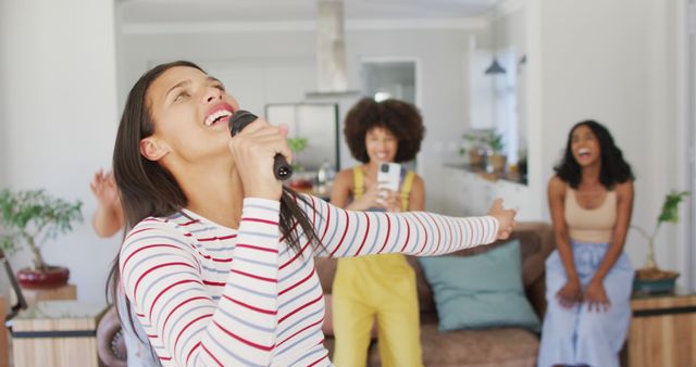 Group of young women having a lively karaoke session in living room, singing with microphone, and enjoying quality time together. Perfect for concepts like friendship, entertainment, youth culture, and indoor activities.