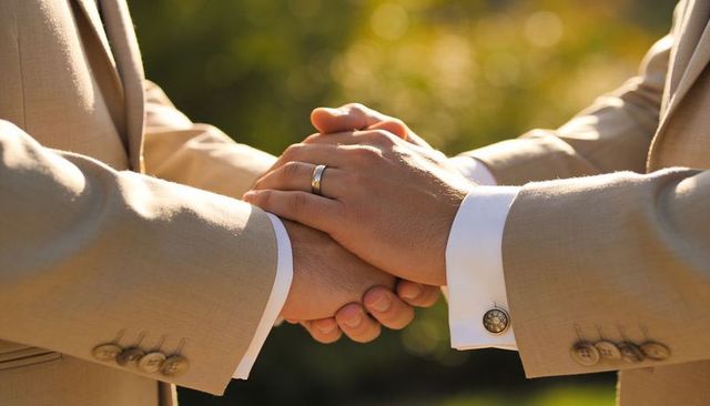 Two grooms clasping hands in beige suits with wedding band during sunlit outdoor ceremony