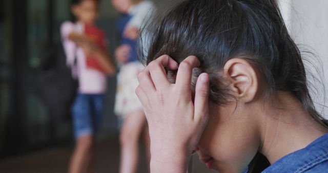 A young girl holds her head in her hands while being bullied by peers in the background. This visual can be used in campaigns and educational materials addressing bullying, mental health in schools, emotional well-being among children, and anti-bullying initiatives.