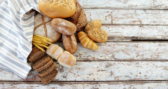 Assorted fresh bread rolls and a bundle of wheat are arranged on a rustic wooden table, with copy space. A variety of textures and shapes offer a visual feast, symbolizing abundance and the simple pleasures of bakery items.