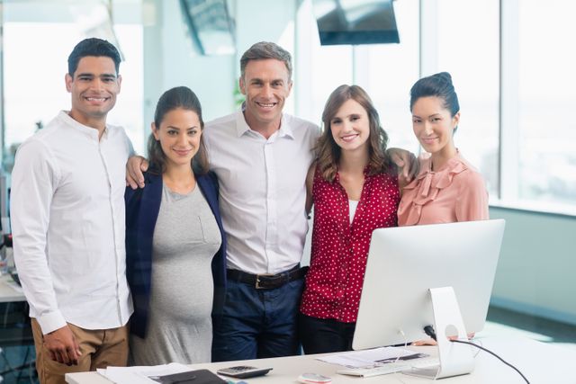 Group of diverse business colleagues standing together at a desk in a modern office, smiling and showing camaraderie. Ideal for use in corporate websites, team-building presentations, and business-related marketing materials to convey teamwork, collaboration, and a positive work environment.