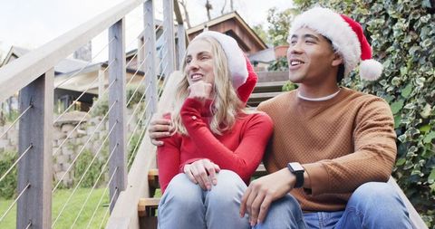 Festive couple relaxing on outdoor steps wearing santa hats and cozy sweaters, smiling