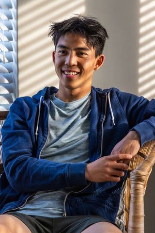Young asian man smiling while sitting in sunlit rattan chair wearing navy hoodie