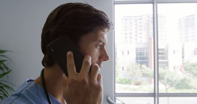 Medical professional listening intently to a phone call while standing by a large window in an office environment. Useful for illustrating concepts related to medical consultations, professional communication, patient care, and modern healthcare settings.