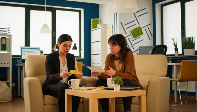 Two women collaborating and reviewing papers while sorting sticky notes in modern office lounge