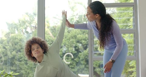 Fitness trainer guiding client holding side-plank beside sunlit window in home workout