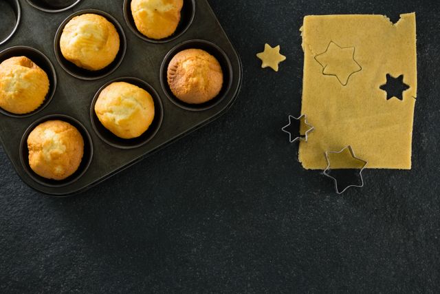 Freshly baked cupcakes in a baking tray next to star-shaped dough and cookie cutters on a dark surface. Ideal for use in baking blogs, recipe websites, or cooking magazines to illustrate homemade baking processes and dessert preparation.