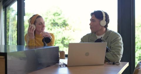 Podcasting duo recording conversation in bright home studio with headphones and laptop