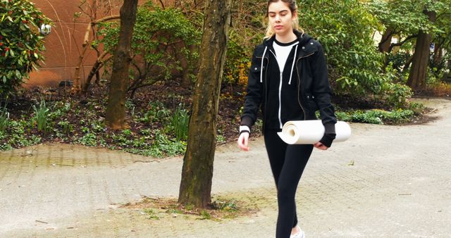 Young woman wearing casual clothes carrying a yoga mat while walking in an outdoor park. The peaceful garden scenery can be used for illustrating themes such as fitness, exercise, wellness, outdoor activities, and healthy lifestyles. This can also be used for promotions of yoga classes or fitness events.