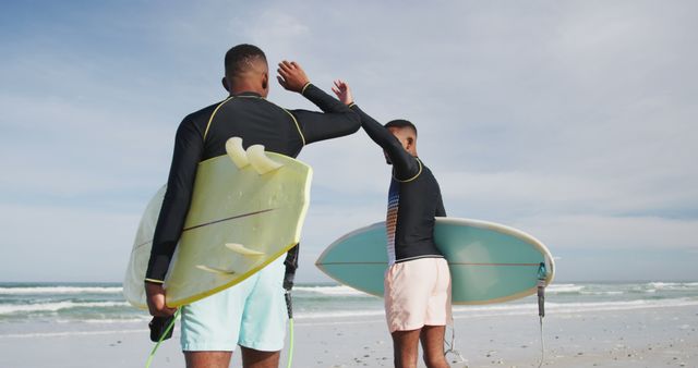 Two friends high-fiving on beach while holding surfboards. Ideal for summer, friendship, and outdoor sports themes. Perfect for promotional materials, advertisements, and social media posts related to surf culture and vacation.