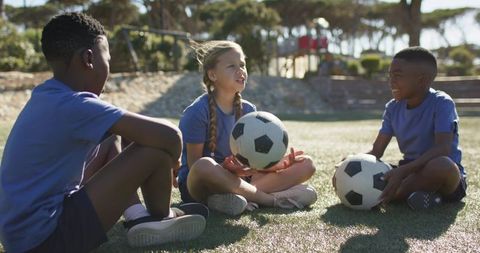 Multiracial youth soccer team sitting on sunny field holding balls and laughing