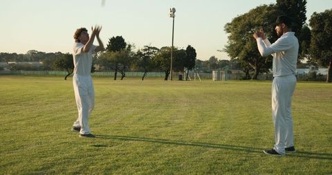 Two cricketers practicing catching drill on sunlit grass field wearing white uniforms