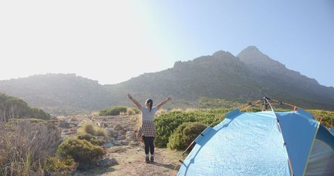 Woman Exploring Mountain Landscape Near Blue Tent in Sunlit Setting