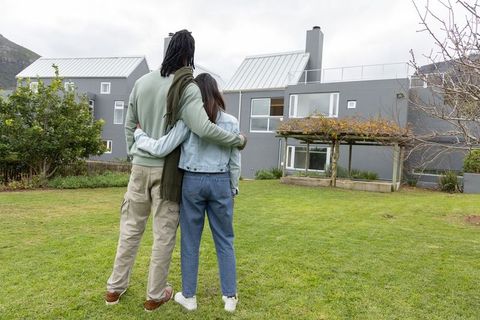 Couple embracing while viewing modern gray two-story house with pergola and spacious lawn