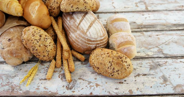 Assorted variety of freshly baked breads and rolls displayed on rustic wooden table. There are whole grain rolls, artisan loaves and crispy breadsticks arranged in a scattered fashion. Ideal for use in food blogs, recipe websites, bakery advertisements and menus that emphasize homemade and artisanal bakery products.