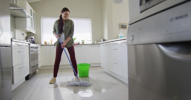Woman mopping floor in modern, white kitchen, showing the regular household cleaning routine. Great for advertisements about cleaning products, home maintenance, or lifestyle blogs promoting cleanliness and orderliness.