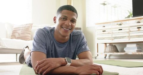 African american man relaxing on green yoga mat in sunlit living room wearing smartwatch