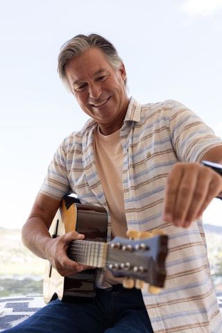 Smiling senior man tuning acoustic guitar on sunny patio enjoying relaxed outdoor music