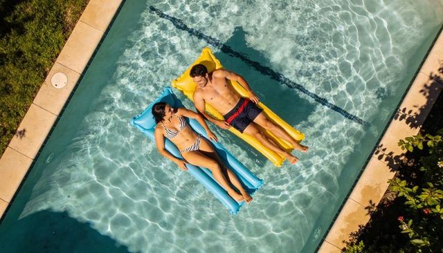 Couple floating on colorful inflatable loungers in sunny backyard pool, relaxing together