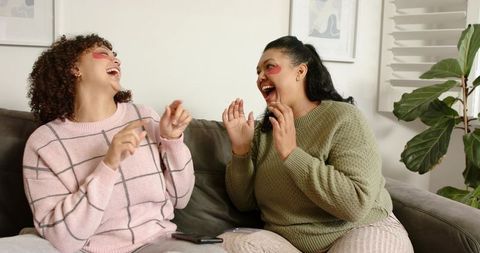 Two Friends Laughing on Couch Sharing Self-Care Moment with Under-Eye Patches and Smartphones