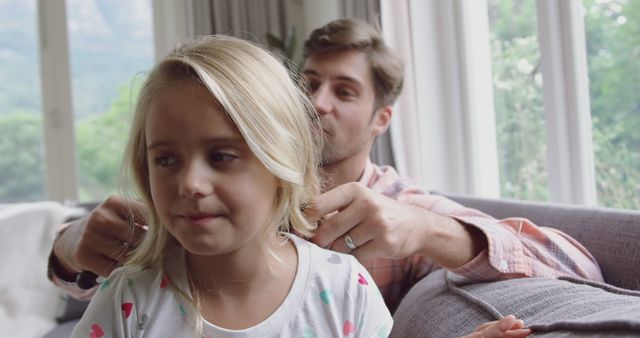 Father bonding with his young daughter by braiding her hair while sitting at home. This image can be used in contexts related to parenting, family relationships, father-daughter activities, and home life. It conveys love, care, and the importance of small everyday moments in building strong family ties.