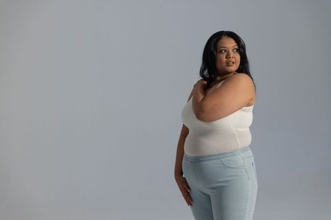 Confident plus-size woman posing in white tank and high-waist jeans on neutral backdrop