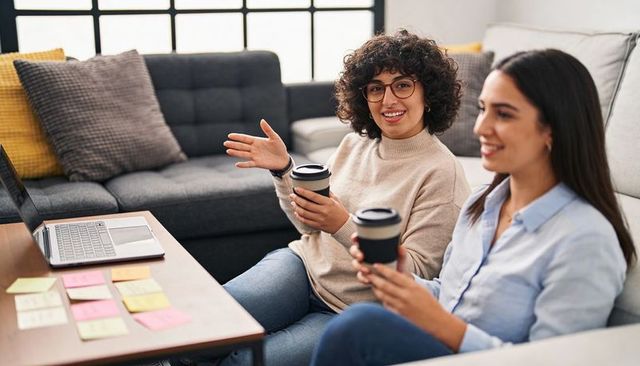 Two women collaborating over coffee on sofa with laptop and sticky notes