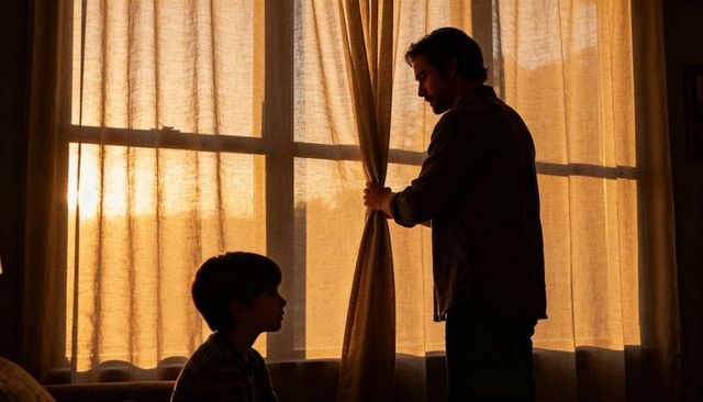 Man pulling curtain while boy watching golden hour light through living room window
