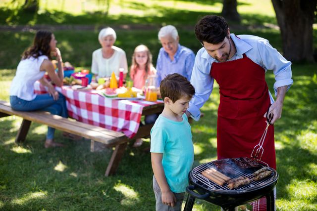 Father and son grilling food together in a park while the rest of the family enjoys a picnic at a table in the background. Ideal for use in advertisements, family-oriented content, summer activity promotions, and articles about family bonding and outdoor leisure activities.