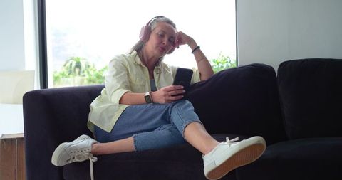 Mature woman relaxing on couch listening with headphones while using smartphone