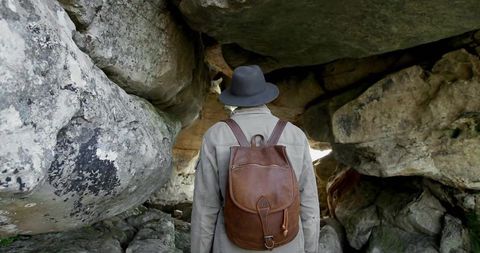 Backpacker exploring rocky overhang peering into crevice with leather backpack and hat